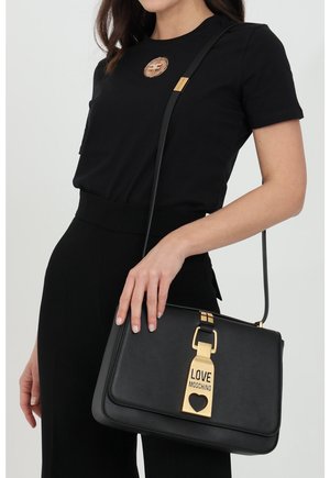 Femme vêtue d'une tenue noire tenant un sac à main Love Moschino noir avec fermeture dorée et découpe en forme de cœur, bandoulière visible.