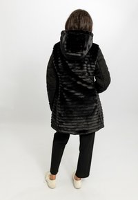 Zwarte nepbontjas met een capuchon, met een ribbeltextuur op de rug en gladde mouwen, gecombineerd met zwarte broeken en witte schoenen.