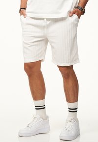 Weiße, strukturierte Shorts mit vertikalen Streifen, elastischem Bund, Eingrifftaschen, kombiniert mit weißen Sneakers und Socken mit schwarzen Streifen.