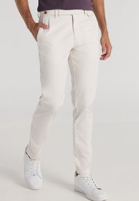 Chinos beige claro hechos de una tela suave, con corte recto, dos bolsillos laterales y cierre de cremallera frontal. Llevados con zapatillas blancas.