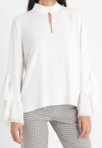 Blusa bianca a maniche lunghe con colletto alto e scollatura a chiave. Presenta maniche a campana e un tessuto liscio e leggero.