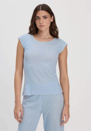 Frau mit schulterlangem braunem Haar, die ein hellblaues, geripptes Kurzarmoberteil und passende Hosen mit dezentem Blumenmuster trägt.