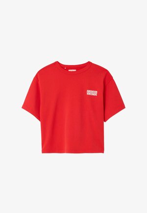 Dieťa s kučeravými vlasmi, oblečené v príliš veľkej červenej košeli, modrých šortkách a béžových nazúvacích topánkach, stojace na jednoduchom bielom pozadí.