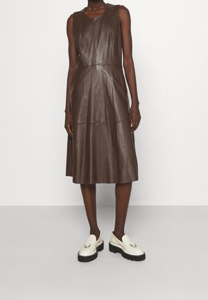 Modelka w ciemnobrązowej, bezrękawnikowej sukience midi ze skóry, zestawiona z białymi loafersami na platformie, na tle jasnego, jednolitego tła.