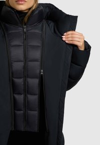 Geacă matlasată neagră cu căptușeală interioară, două fermoare și glugă. Prezintă un material exterior neted și un design structurat, cu umplutură .