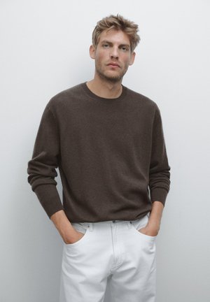 Blond man wearing a dark brown sweater and white pants with hands in pockets, standing against a plain light gray background.