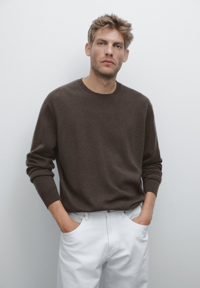 Uomo biondo che indossa un maglione marrone scuro e pantaloni bianchi, con le mani in tasca, in piedi contro uno sfondo grigio chiaro uniforme.