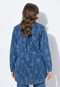 Camicia di denim con piega posteriore, caratterizzata da un motivo astratto blu e maniche lunghe. I dettagli includono polsini con bottoni e un orlo arrotondato.