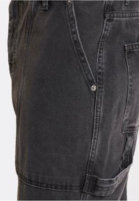 Pantalones de mezclilla negros tipo overol con un bolsillo lateral, botones de metal y detalles de costura visibles. La tela tiene una textura suave con un ligero brillo.