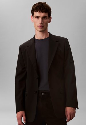 Sort blazer med to knapper, hakkede revers og tilpasset pasform, båret over en mørk t-shirt. Enkel design med glat tekstur.