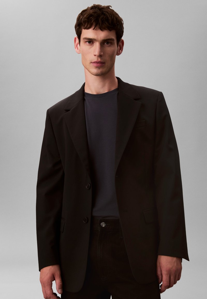 Schwarzer Blazer mit zwei Knöpfen, geschlitzten Revers und taillierter Passform, getragen über einem dunklen T-Shirt. Einfaches Design mit glatter Textur.