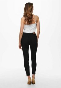 Mujer con cabello largo castaño, vistiendo una camiseta sin mangas blanca, jeans ajustados negros y tacones altos beige, de pie de espaldas sobre un fondo blanco.