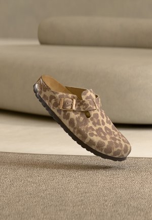 Single leopard print slip-on shoe with buckle detail, floating above a textured beige carpet and neutral sofa background.