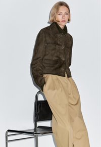 Hnědá semišová bunda se zapínáním na knoflíky vpředu, dvěma kapsami a krátkým střihem. Kombinováno s béžovými plisovanými kalhotami.