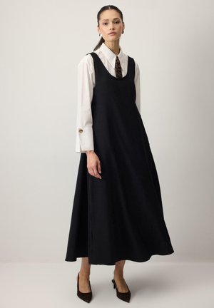 Femme portant une robe noire sans manches jusqu'aux chevilles sur une chemise blanche boutonnée avec une cravate à imprimé tigre, assortie à des talons pointus foncés, debout.