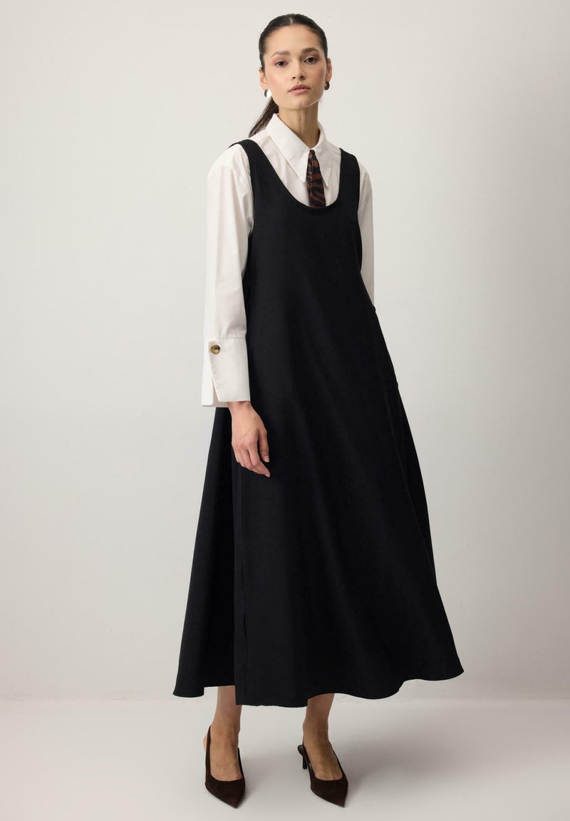 Kobieta ubrana w czarną, bezrękawnikową sukienkę do kostek na białej, zapinanej na guziki koszuli z krawatem w paski tygrysa, w połączeniu z ciemnymi, szpiczastymi obcasami, stojąca.