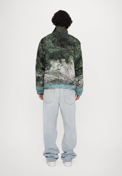 Padded jacket featuring a nature print with green trees and rocks; high collar, wide sleeves, and blue accents at the hem. Worn with light jeans.