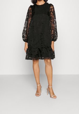 Kobieta ubrana w czarną sukienkę do kolan z kwiatowymi, przezroczystymi długimi rękawami i dołem, zestawioną z srebrnymi sandałami na wysokim obcasie.