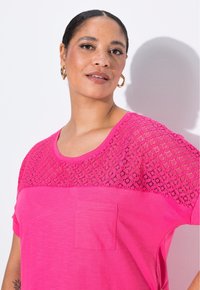 Světle růžové tričko s krátkým rukávem, které má krajkovanou část na ramenou a malou přední kapsu; měkká tkanina s volným střihem.