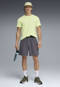 Νεαρός άνδρας που στέκεται φορώντας ανοιχτό κίτρινο μπλουζάκι, γκρι σορτς, ελιά πράσινα αθλητικά παπούτσια, άσπρες κάλτσες Puma, μπεζ καπέλο, κρατώντας ένα διαφανές μπουκάλι νερού Puma.