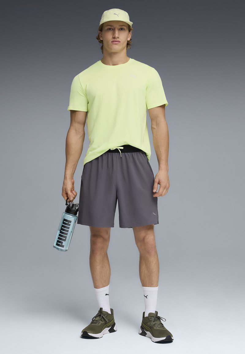 Νεαρός άνδρας που στέκεται φορώντας ανοιχτό κίτρινο μπλουζάκι, γκρι σορτς, ελιά πράσινα αθλητικά παπούτσια, άσπρες κάλτσες Puma, μπεζ καπέλο, κρατώντας ένα διαφανές μπουκάλι νερού Puma.