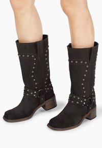 Stivali neri in suede alti fino al ginocchio con borchie argentate lungo il lato e una fascia con fibbia, caratterizzati da un tacco in legno stratificato.