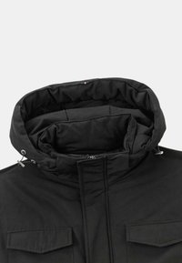 Veste noire avec un col montant, fermeture à glissière et deux poches avant. Le matériau semble lisse avec une texture intérieure douce et rembourrée.