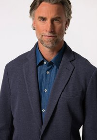 Granatowa marynarka o fakturze z wyciętymi klapami, zestawiona z dżinsową koszulą z guzikami. Zauważalny detal kieszeni na marynarce.