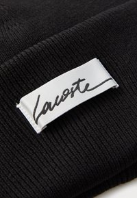 Sort ribbet strikket stof med et hvidt rektangulært mærke, der har det håndskrevne brandnavn "Lauoste" i sort blæk.