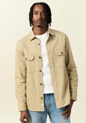 Une chemise boutonning kaki présente deux poches poitrine, des boutons marron et une texture lisse, portée par-dessus une chemise blanche à côtes et un jean bleu.