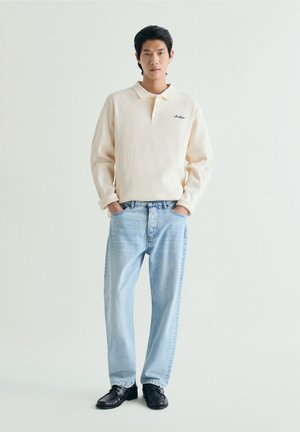 Joven de pie con las manos en los bolsillos de unos vaqueros azul claro, vistiendo una camisa polo crema de manga larga y zapatos negros sobre un fondo liso.