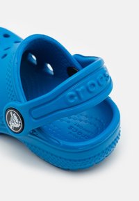 Zilas Crocs sandales, kas izgatavotas no viegla sintētiska materiāla ar ventilācijas atverēm, mīkstu zolīti un ar zīmola jostu pāri augšai.
