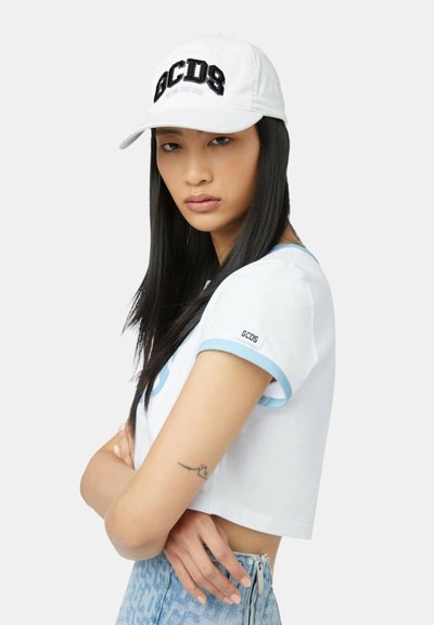 Młoda kobieta z długimi czarnymi włosami, nosząca białą czapkę GCDS, krótki biały T-shirt z niebieską lamówką oraz jasnoniebieskie wzorzyste spodnie, z założonymi skrzyżowanymi ramionami.