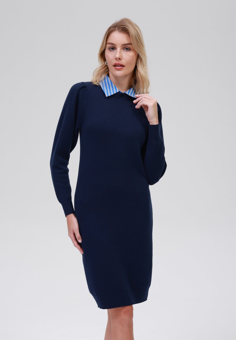 Robe tricotée bleu marine avec des manches longues et une silhouette ajustée ; présente un col rayé bleu clair. Texture lisse avec des accents côtelés aux poignets et à l'ourlet.