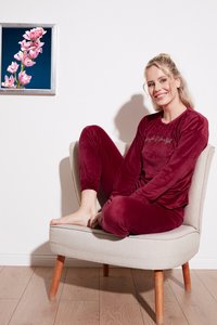 Burgundijska velur pidžama s dugim rukavima i suženim hlačama, s natpisom "Jednostavno i Lijepo" u zlatnoj boji. Svijetla drvena stolica i cvjetna umjetnost.