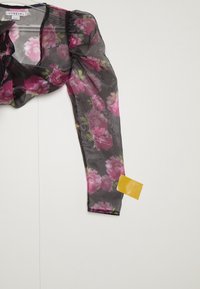 Genomskinlig svart blus med rosa blommönster, puffärmar och en gul tagg fäst vid vänster manschett. Lätt material, elegant design.