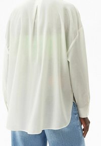 White, semi-transparent long-sleeve shirt featuring a back vent, relaxed fit, and buttoned cuffs. Paired with light blue denim jeans.