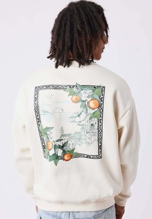 Personne aux cheveux bouclés et foncés porte un sweat-shirt blanc avec un motif représentant un village côtier pittoresque et une branche d’orange dans le dos.