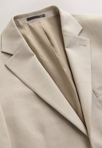 Blazer beige avec col à cran, boutonnière simple et doublure intérieure, posé à plat sur une surface claire.