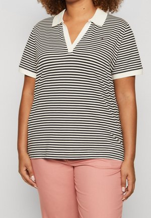 Kobieta ubrana w czarno-białą, poziomo prążkowaną, krótko rękawową koszulkę polo z białym kołnierzykiem i różowe spodnie, stojąca na jednolitym tle.