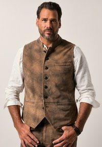 Chaleco de cuero marrón con un alto cuello, seis botones negros y un bolsillo en el pecho. Lleva puesto una camisa blanca de manga larga. Textura suave.