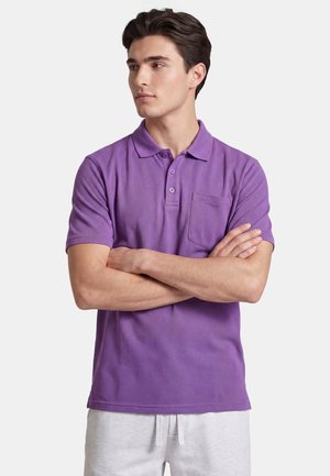Junger Mann mit dunklen Haaren, der ein lila Polohemd mit Tasche und helle Hosen trägt, verschränkte Arme, schaut zur Seite.
