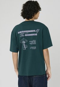 T-shirt en coton vert à manches courtes. Le dos comporte un grand texte "ÉVEIL DES SENS" et des graphismes de champignon en blanc et violet.