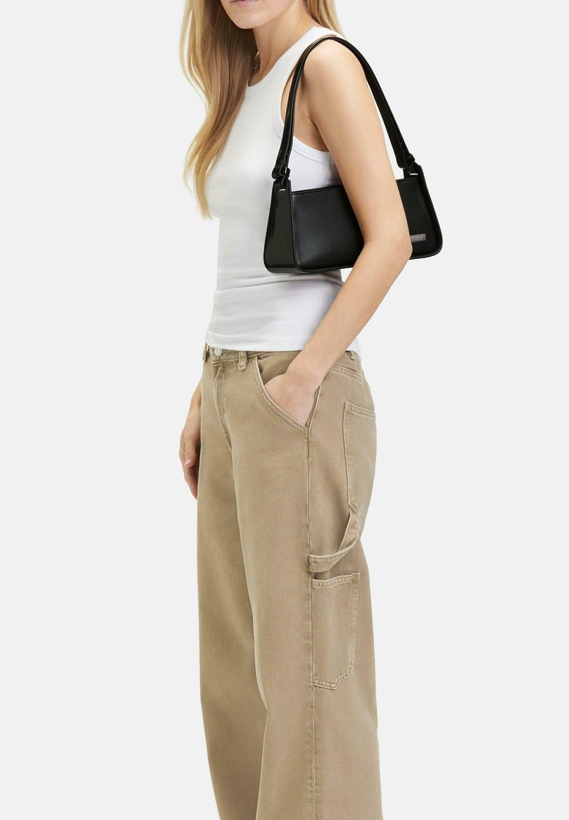 Borsa a mano nera in pelle con un design strutturato e una tracolla corta, posizionata accanto a una persona che indossa una canotta bianca e pantaloni cargo beige.