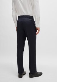 Jūriniai klasikiniai kelnės su pritaikytu siluetu, lygiu paviršiumi, dviem galiniais kišenėmis ir diržo kilpos. Dėvimos su juodais batais, matomos iš nugaros.