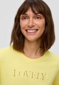 T-shirt en coton jaune avec un design texturé "LOVELY". Le haut a un col rond et une texture lisse.