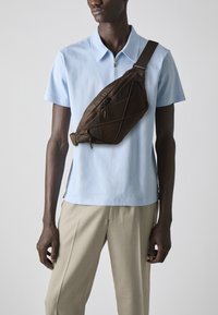 Brown nylon sling bag with geometric design, diagonal seams, and a zippered pocket. Worn across a light blue polo shirt and beige trousers.