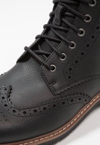 Stivale in pelle nera con dettagli perforati, punta rotonda e occhielli metallici. Presenta una suola cucita di colore marrone e una superficie texturizzata.