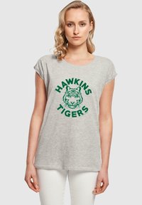 Pelēks kokvilnas T-krekls ar sarullētām piedurknēm, rotāts ar zaļu tīģera grafiku un tekstu "HAWKINS TIGERS" apļveida dizainā.