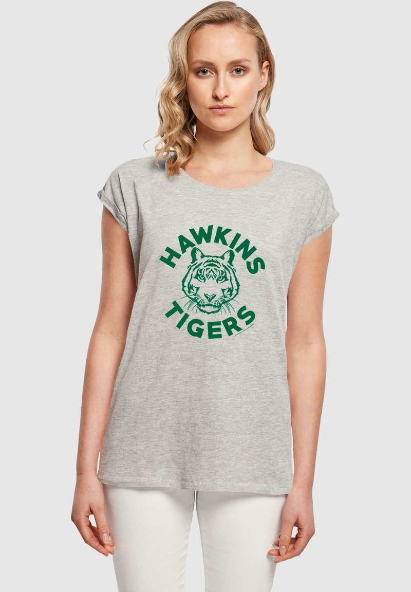 Pelēks kokvilnas T-krekls ar sarullētām piedurknēm, rotāts ar zaļu tīģera grafiku un tekstu "HAWKINS TIGERS" apļveida dizainā.